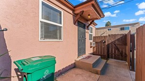 Un edificio residencial con fachada color crema, una puerta de madera y un contenedor verde para reciclaje.