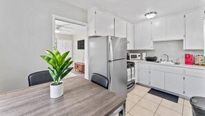 Una cocina moderna con armarios blancos, un refrigerador de acero inoxidable, un microondas y un lavamanos. Hay una mesa de comedor con sillas y una planta en maceta.