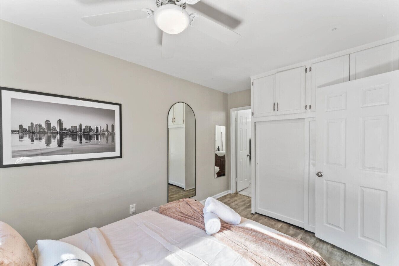Un dormitorio con cama, ventilador de techo, cuadro enmarco con vista de la ciudad, espejo de cuerpo entero y armario blanco.