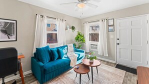 Una sala de estar moderna con un sofá azul, un ventilador de techo y vistas a la calle a través de las ventanas.