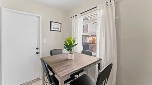 Un comedor con una mesa de madera, cuatro sillas, una planta en maceta, un cuadro en la pared y una ventana con cortinas blancas.