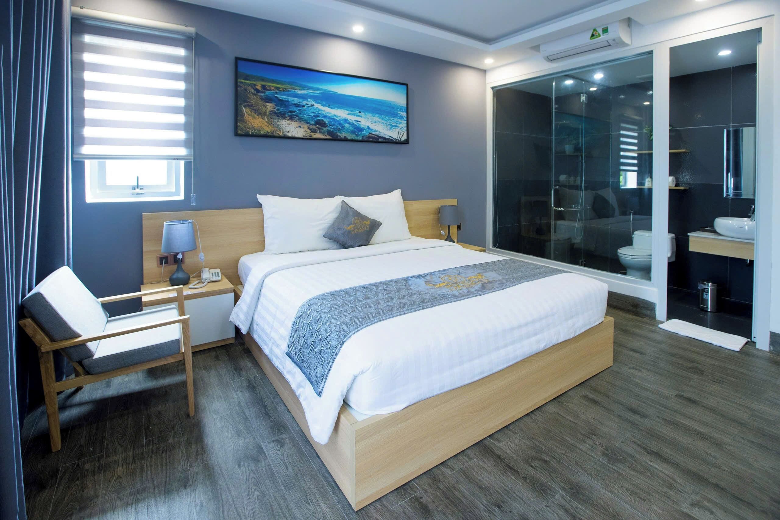 A modern hotel room with a large bed, a bedside table with a lamp, a chair, a painting on the wall, and a glass shower enclosure.