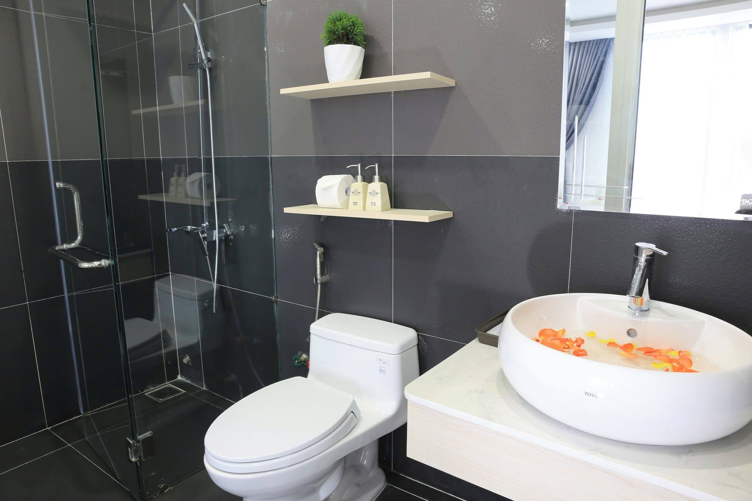 A modern bathroom with a glass-enclosed shower, a white toilet, a white sink with orange flower petals, and a small potted plant on a shelf.