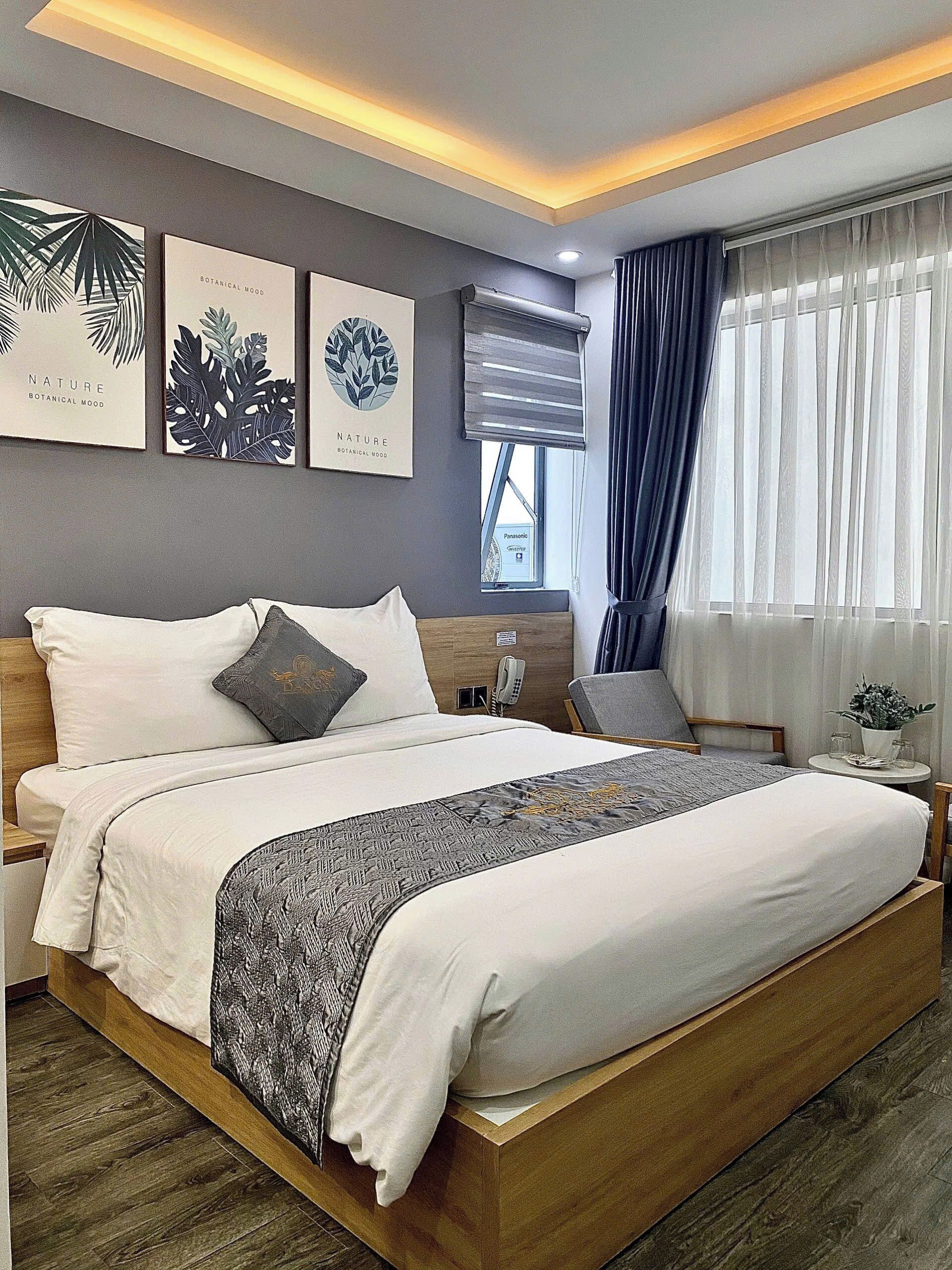 A modern hotel room with a large bed, a bedside table with a lamp, and a view of the outdoors through a window with curtains.