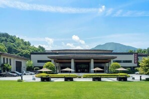 Un edificio moderno con acceso cubierto, estacionamiento y montañas al fondo.