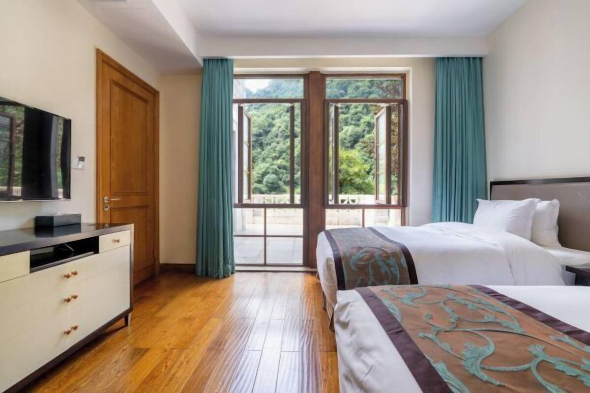 Una camera d'albergo con pavimento in legno, un letto con copriletto decorato, una televisione su un comò e un'ampia finestra con tende.