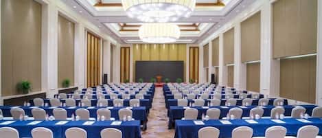 Un salón de conferencias con filas de mesas y sillas, un escenario al frente y un techo alto con candelabros.