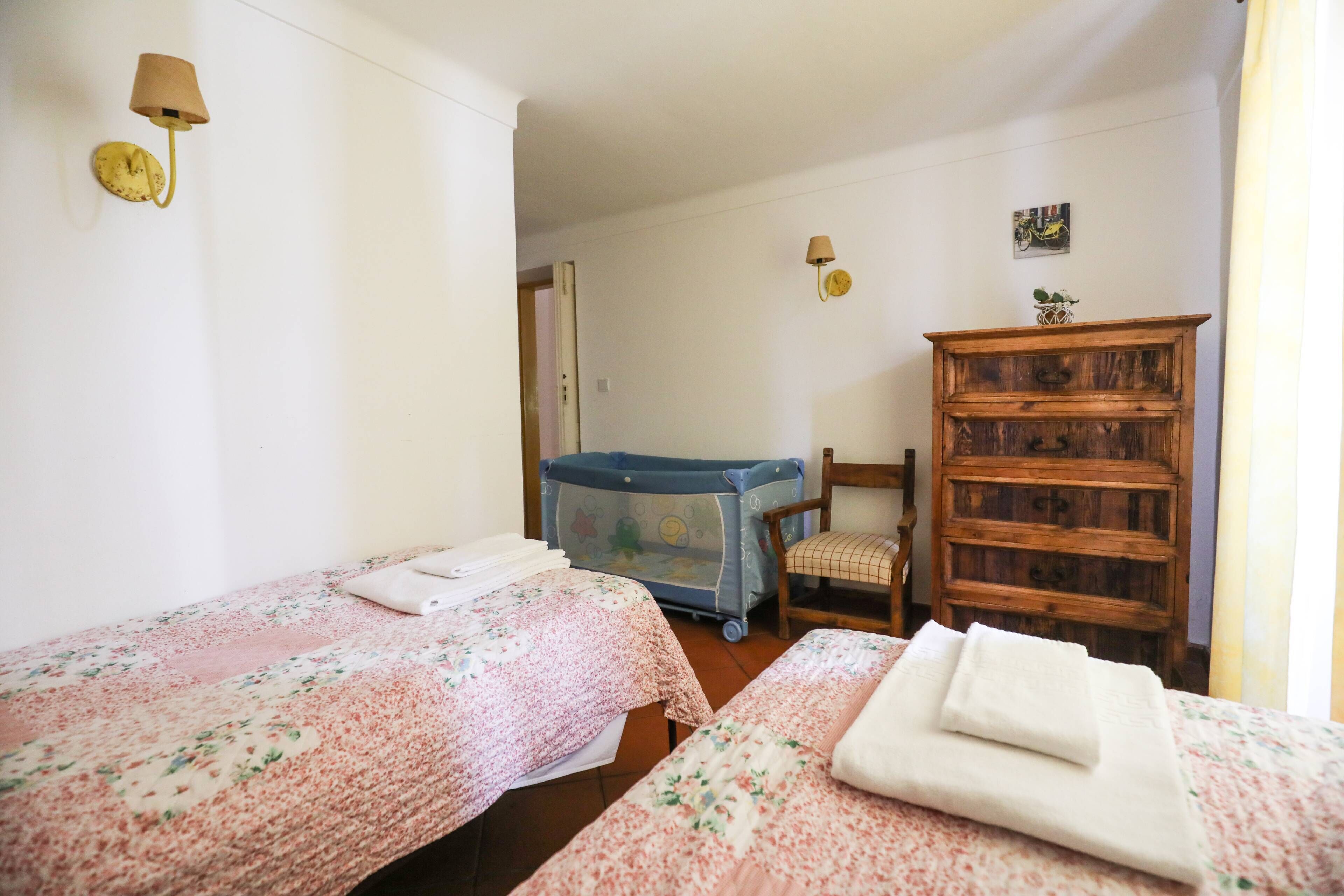 Une chambre avec deux lits, un berceau, une commode en bois et des lampes fixées au mur.