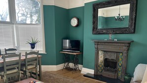 Una sala de estar con paredes color esmeralda, una mesa de comedor de madera, sillas, un televisor sobre un mueble, una chimenea y un espejo grande.