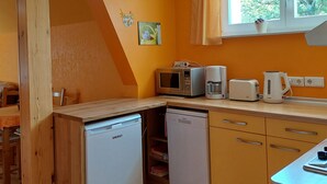 A compact kitchen with a dishwasher, microwave, coffee maker, and toaster. A window with curtains is visible, and a small plant is on the windowsill.