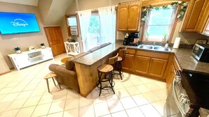 A kitchen with a bar area, wooden cabinets, a tiled floor, a television displaying Disney+, and a window with curtains. - Spacious King Bed Suite at Antique Store (Wakeman)