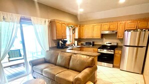 A modern kitchen with wooden cabinets, a stainless steel refrigerator, a built-in oven, and a dining area with a brown leather sofa and a small wooden stool. - Spacious King Bed Suite at Antique Store (Wakeman)
