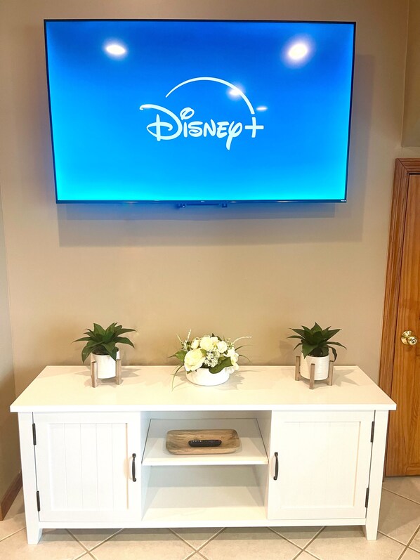 A flat-screen TV mounted on a wall displaying the Disney+ logo. Below the TV, there is a white cabinet with two potted plants on either side and a floral arrangement in the center.