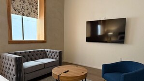 Un salon avec un canapé gris à motifs capitonnés, une table basse ronde en bois, un fauteuil bleu et un téléviseur à écran plat fixé au mur.