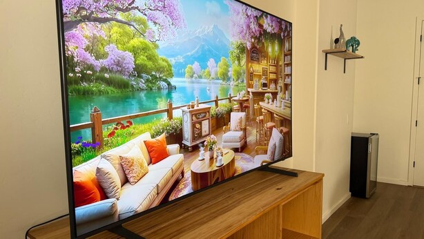 Ein Flachbildfernseher an einem Holzboden, zeigt eine malerische Landschaft mit einem See, Bäumen und einem Gebäude mit Barbereich.