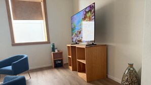 Ein Wohnzimmer mit Fernseher auf einem Holzständer, einem blauen Sessel, einem kleinen Tisch mit Wasserkocher und einem Korkenbehälter.