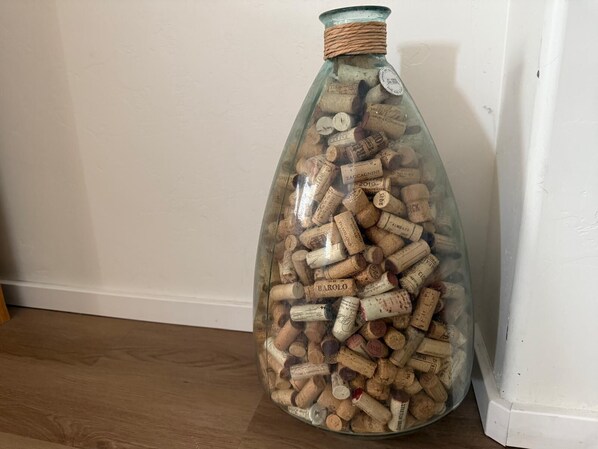 A clear glass bottle filled with wine corks, placed on a wooden floor next to a white wall.