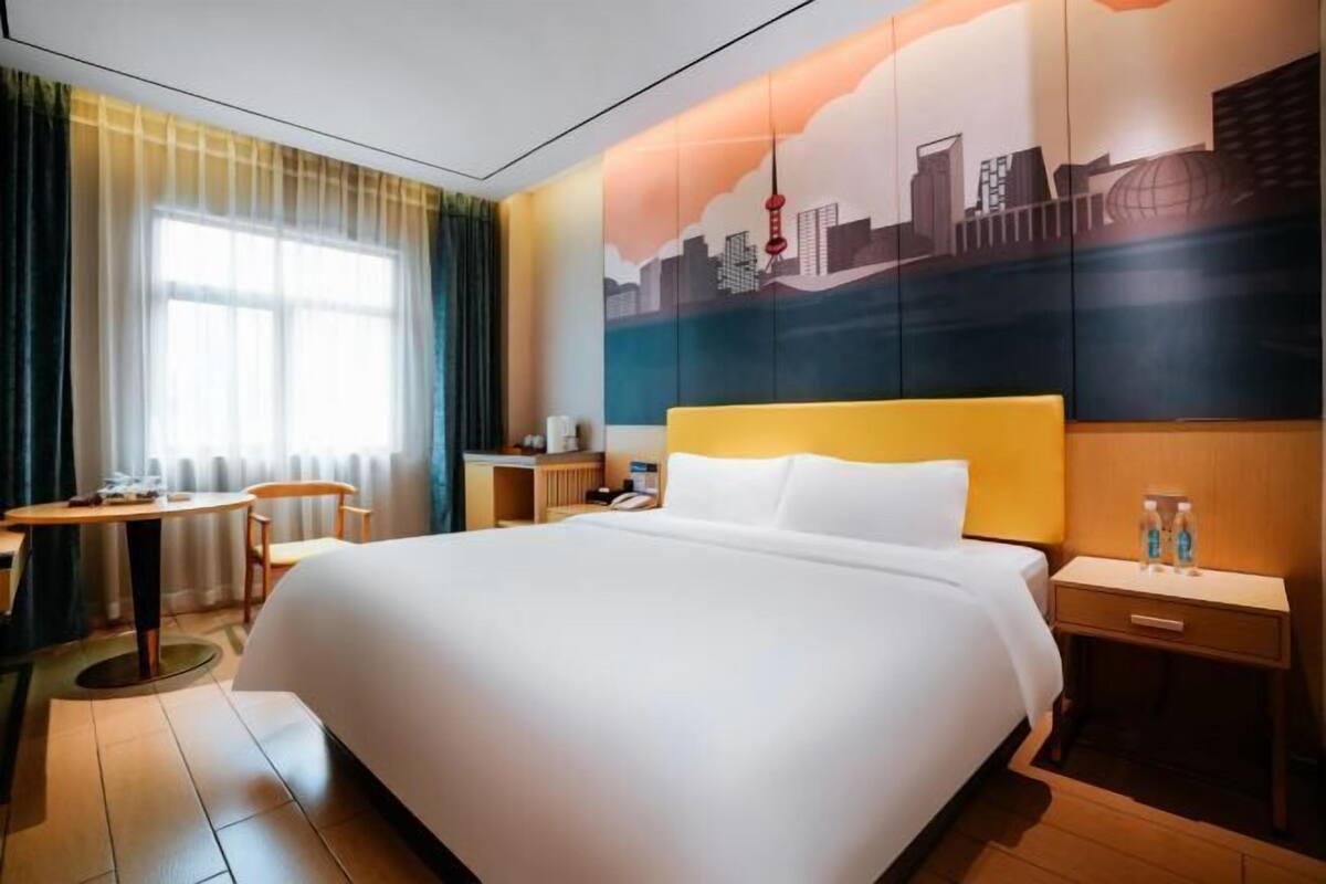 Une chambre d’hôtel avec un grand lit, un bureau, une chaise, une table de chevet et une fresque représentant un paysage urbain sur le mur.