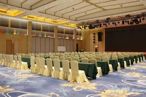 Une salle de conférence avec des rangées de tables et de chaises, une scène avec un système d’éclairage et un grand écran.