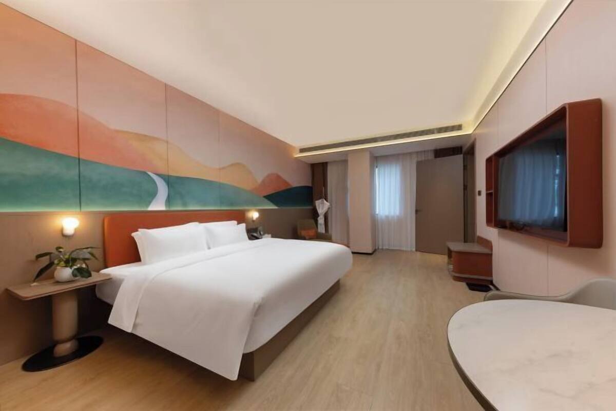 Une chambre d’hôtel équipée d’un lit, d’une table de chevet, d’une lampe, d’une télévision et donnant sur le couloir.