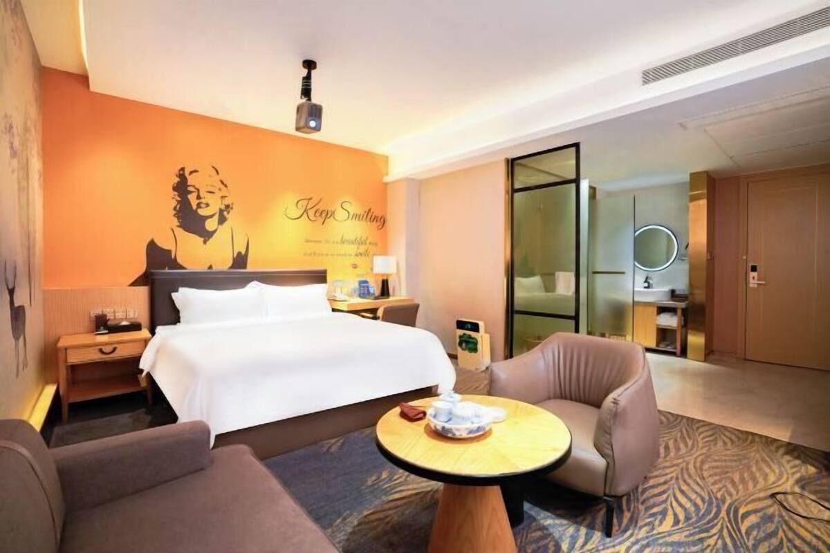 Une chambre d’hôtel comprenant un lit, un bureau, une chaise, une table ronde, un miroir et un mur orné d’une affiche.