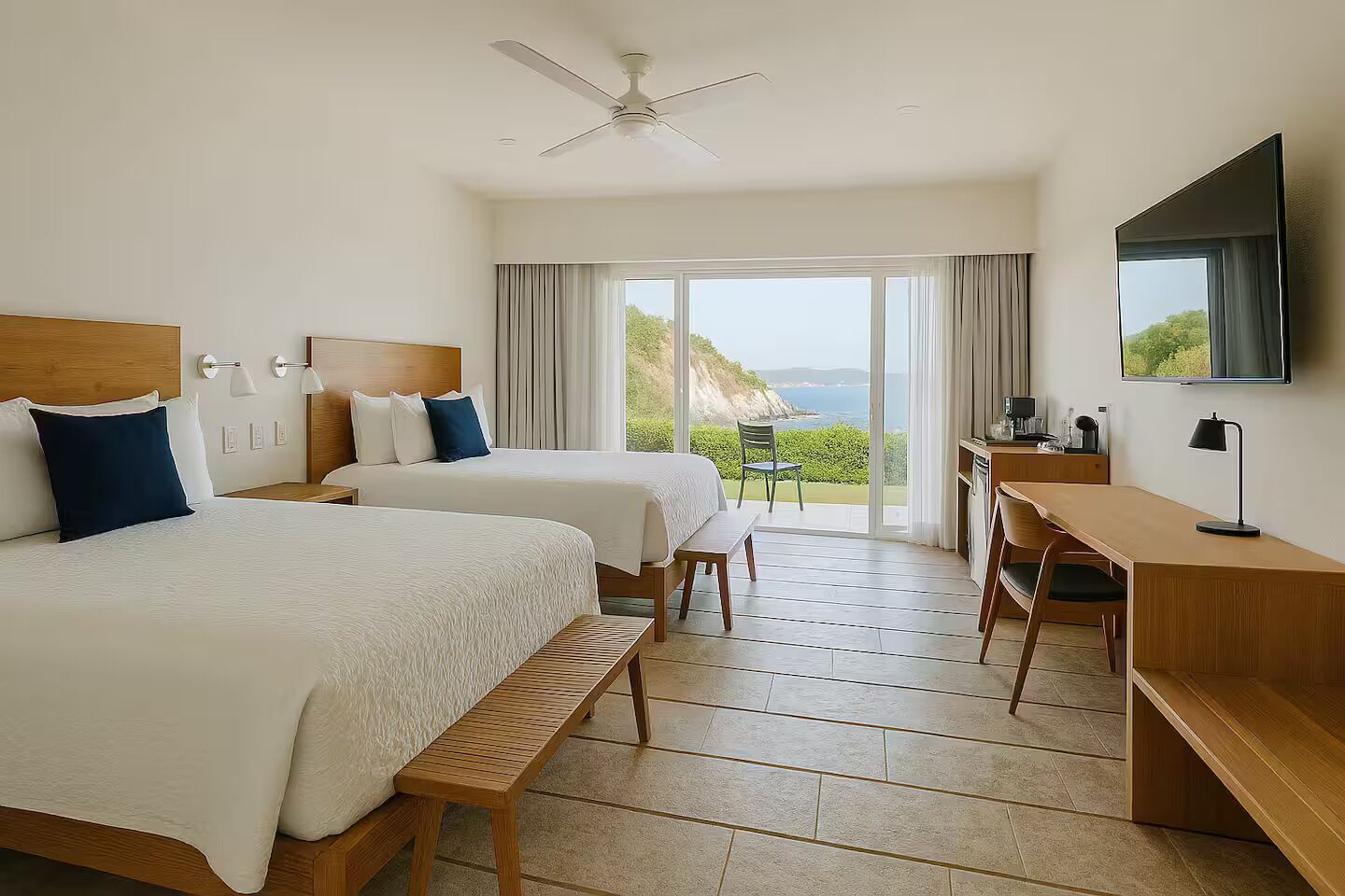 Une chambre d’hôtel avec deux lits, un bureau, une chaise, une télévision et un balcon donnant sur la mer.