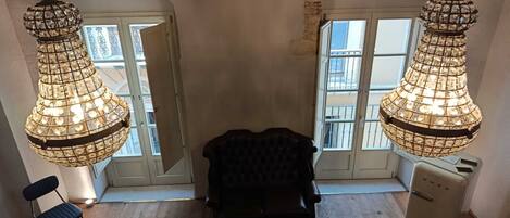 Een kamer met een hoge plafond, twee hangende kristallen kroonluchters, een donkere gestoffeerde stoel, een koelkast in vintage stijl, en een houten vloer.