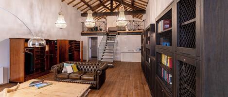 A spacious loft with high ceilings, exposed wooden beams, a leather sofa, a wooden table, and a staircase leading to an upper level.
