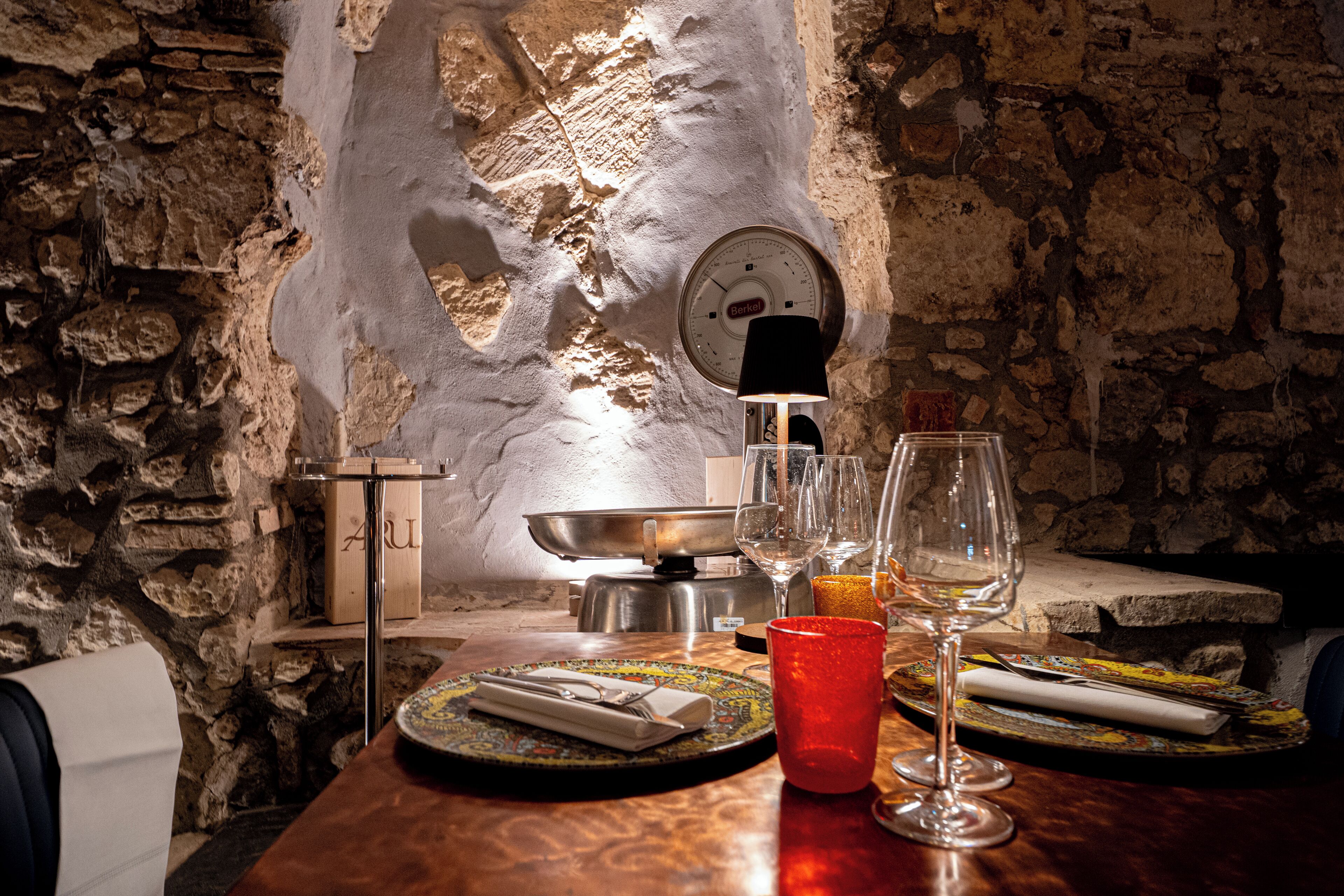 Una mesa de comedor rústica con platos, cubiertos y vasos, frente a una pared de piedra con un reloj.