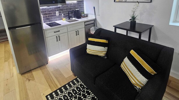 A compact living space with a kitchenette, a black sofa with patterned cushions, a small side table, and a painting on the wall.