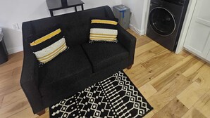 A living room with a black sofa, two cushions, a small table, a vase with flowers, and a painting on the wall.