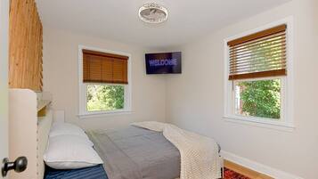 A bedroom with a bed, a TV mounted on the wall, and two windows with blinds.