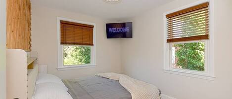 A bedroom with a bed, a TV mounted on the wall, and two windows with blinds.