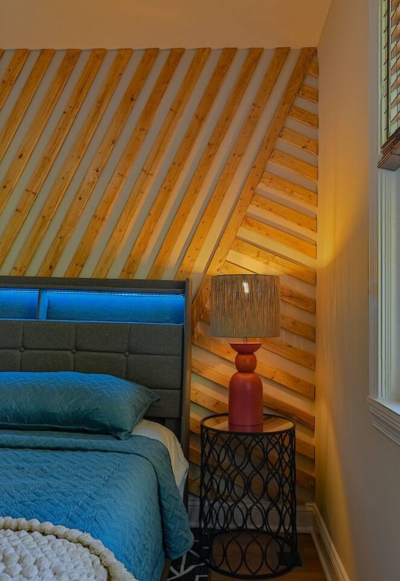 A bedroom with a sloped ceiling, a bed with a blue bedspread, a grey headboard, and a red lamp on a black side table.