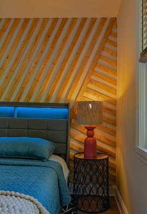 A bedroom with a sloped ceiling, a bed with a blue bedspread, a grey headboard, and a red lamp on a black side table.