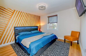 Un dormitorio con cama, una silla, una ventana y un televisor.