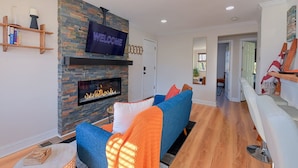 A modern living room with a stone fireplace, a flat-screen TV, and a blue sofa with orange throw pillows. - Treehouse Suite | King BR | Spa bath | Calm Energy (Parkville)