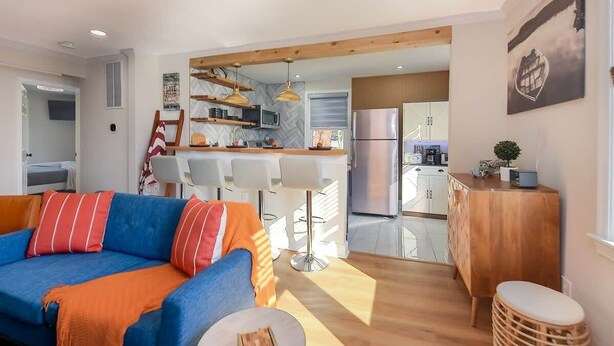 Ein modernes Wohnzimmer mit einem blauen Sofa, einem orangefarbenen Überwurf und einem Essbereich aus Holz mit weißen Stühlen.