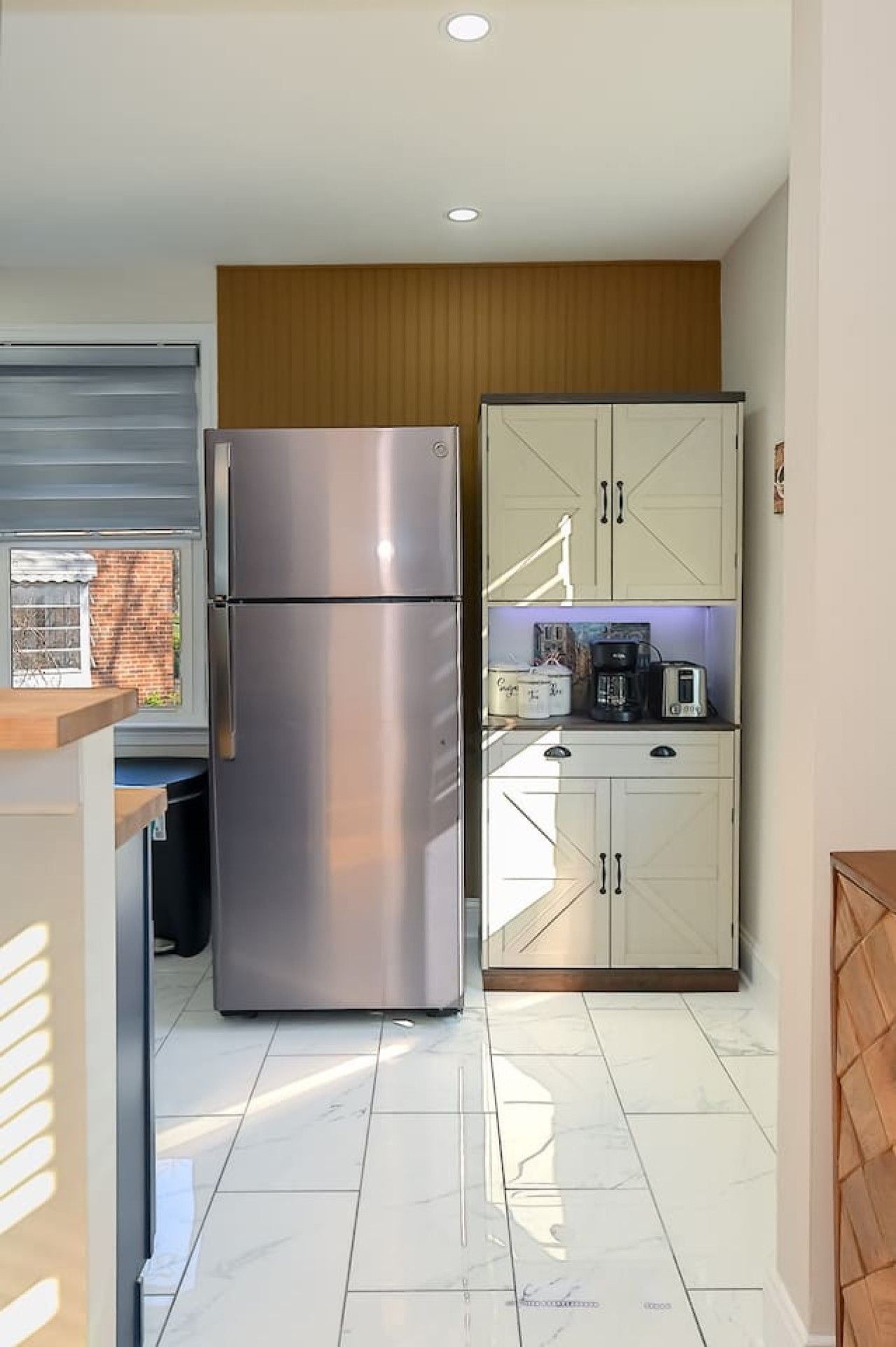 A compact kitchen with a stainless steel refrigerator, a wooden cabinet with kitchen appliances, and a window with a view of a brick building.