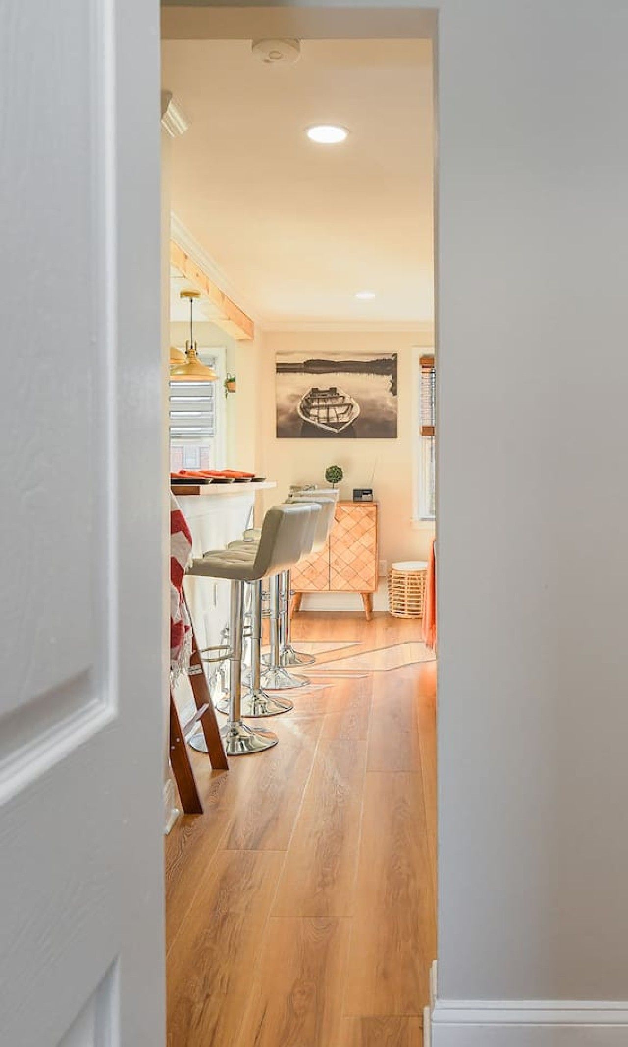 A hallway leading to a dining area with a wooden floor, a bar area with stools, and a kitchen visible in the background.