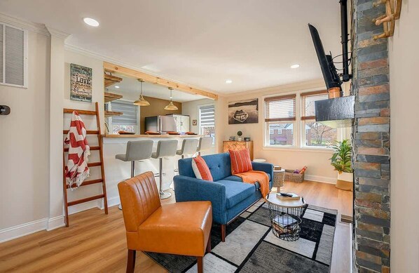 Una sala moderna con un sofá azul, un sillón naranja y una mesa de centro. Se observa una cocina al fondo con un refrigerador y un microondas.