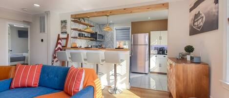 A modern living room with a sofa, a coffee table, and a kitchen area with a refrigerator, microwave, and wooden cabinets.