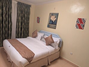 A neatly made bed with a brown throw, a blue headboard, and multiple pillows. There are two wall decorations, one of a flower arrangement and another of a flag. - ECOAIR VILLA (Kigali)