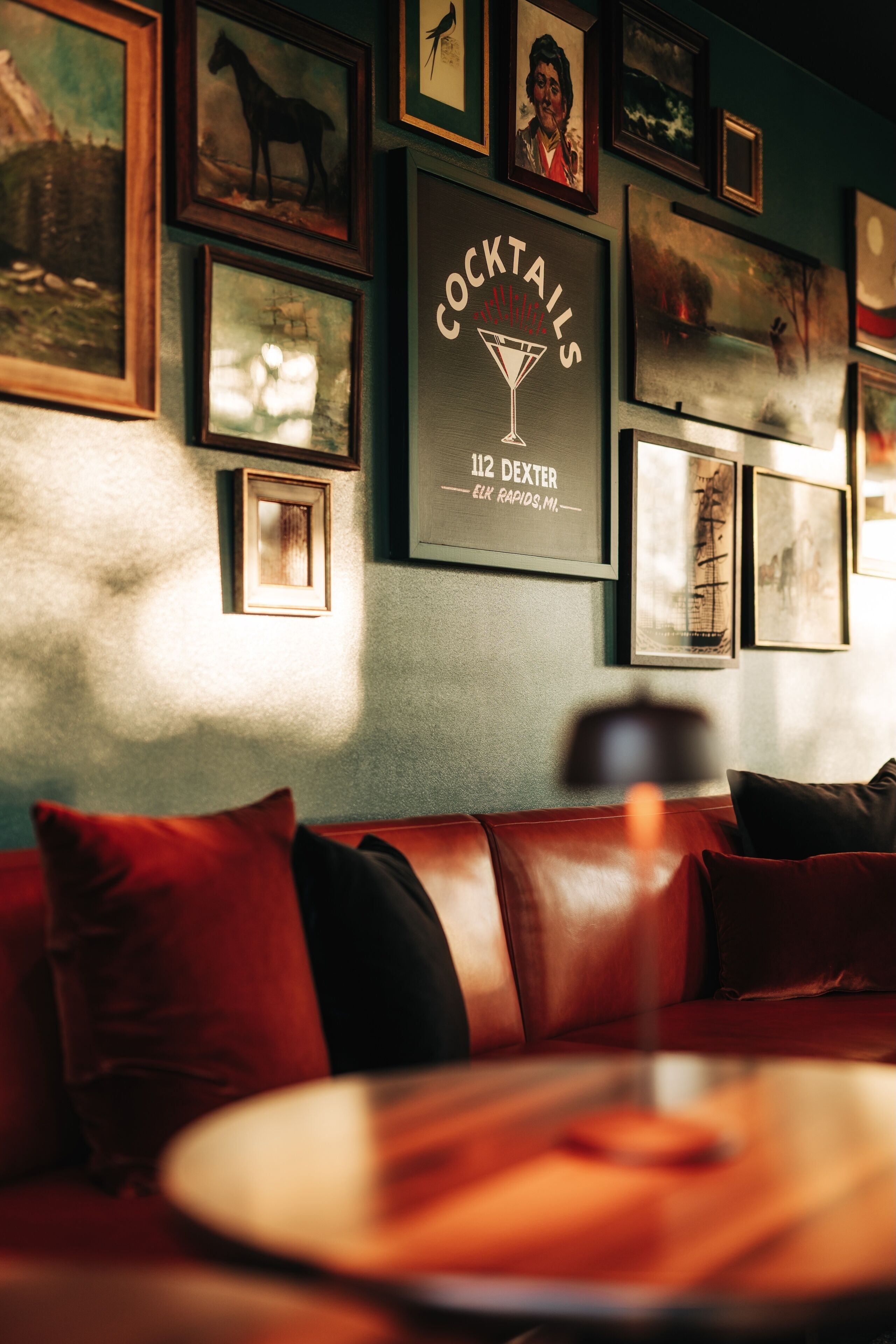 A bar area with framed artwork on the wall, a red leather couch, and a round table with a drink.