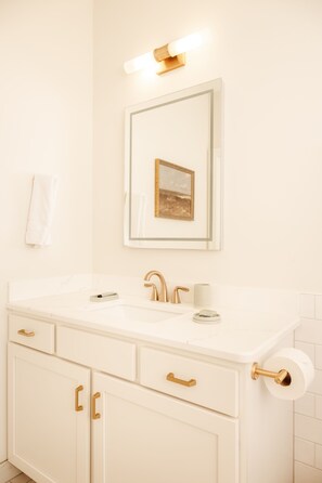 A bathroom with a white vanity, gold fixtures, a framed picture above the mirror, and a towel hanging on the wall. - Dexter Hotel (Elk Rapids)