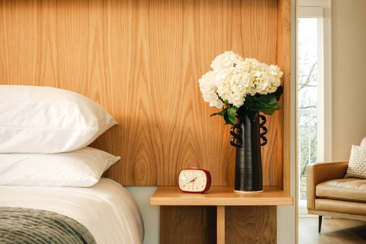 A bedroom with a wooden headboard, a bedside table with a vase of flowers, a clock, and a mirror reflecting a chair and a window.
