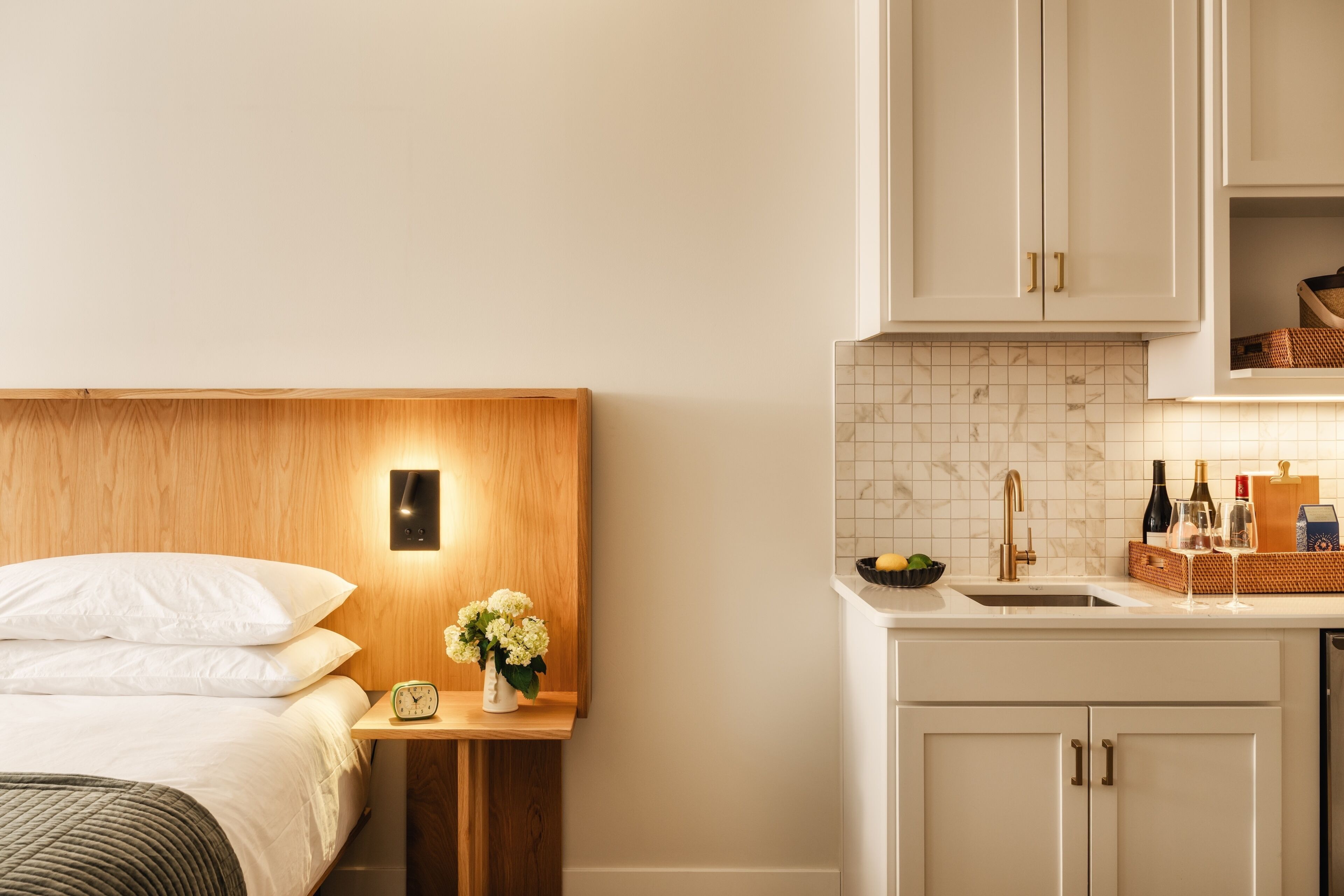 A bedroom with a wooden headboard, a bedside table with a clock and flowers, and a nightstand. Adjacent is a kitchenette with a sink, a fruit bowl, and a basket with bottles and glasses.