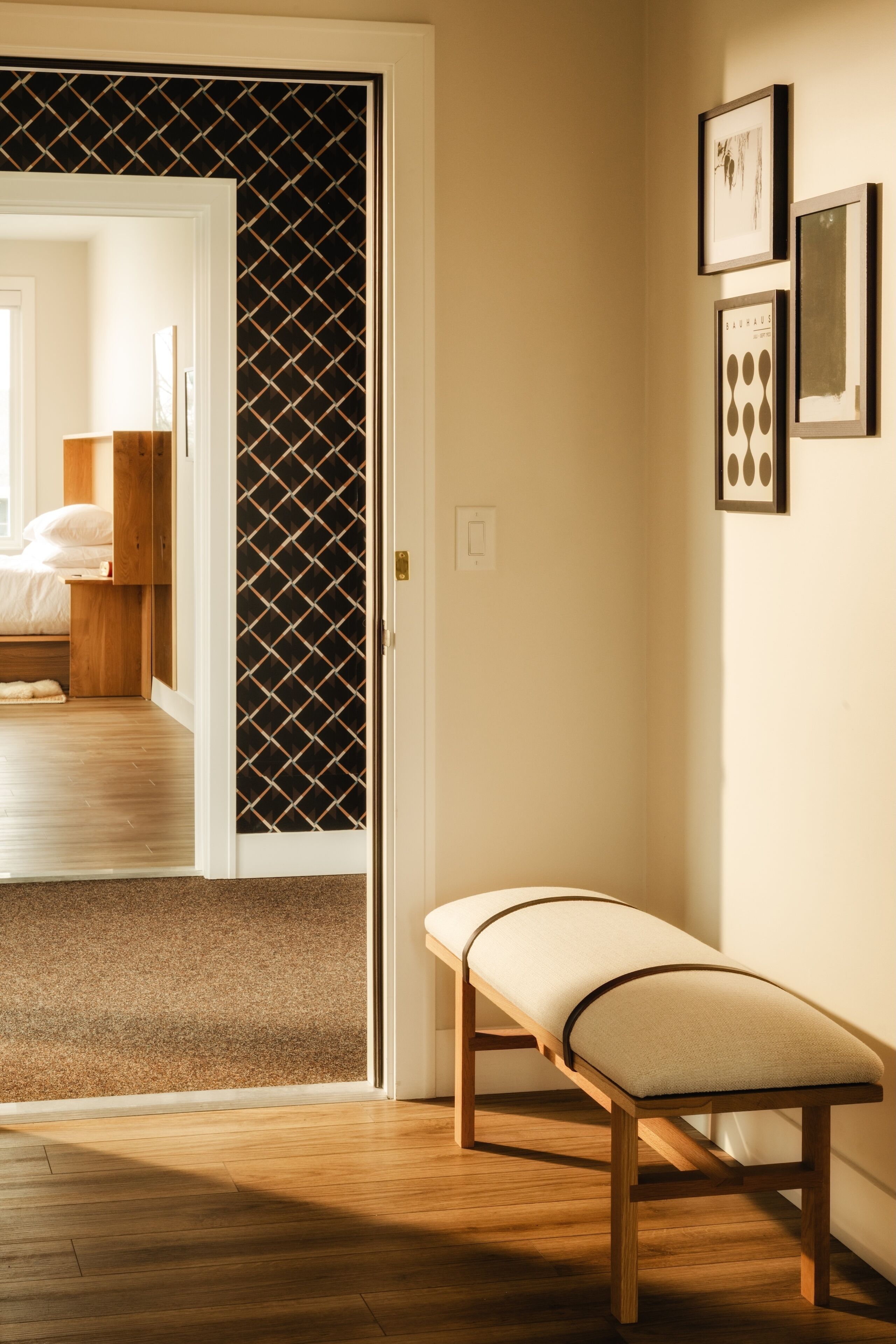 A hallway with a bench, a door leading to a bedroom, and framed artwork on the wall.