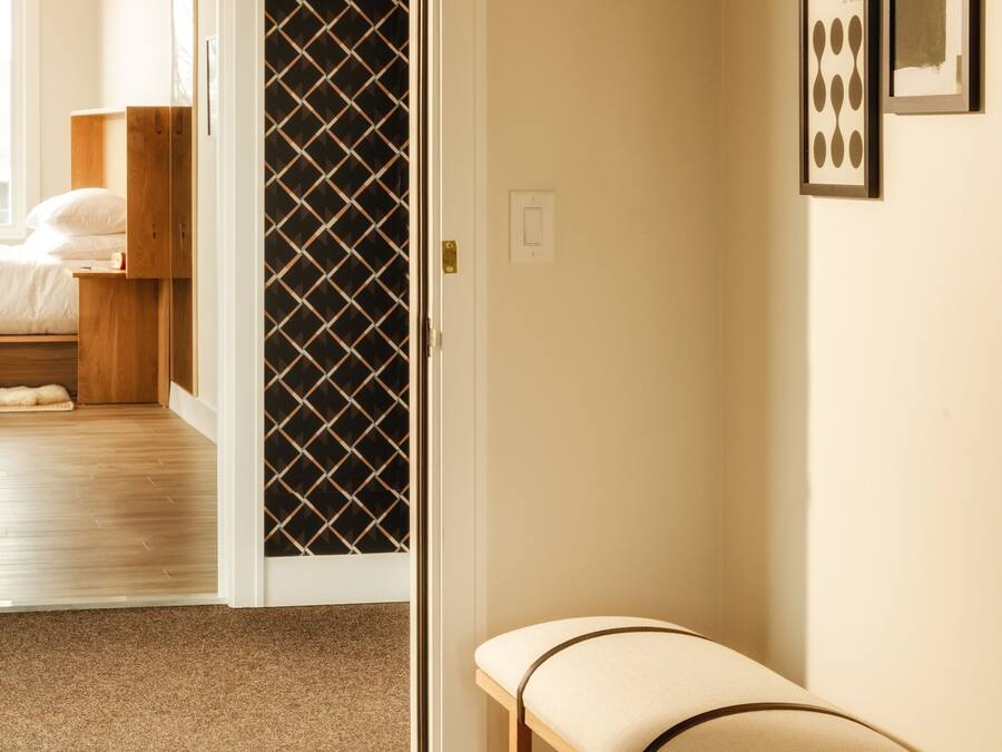 A hallway with a bench, a door leading to a bedroom, and framed artwork on the wall.