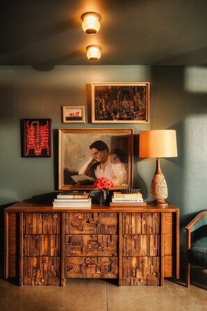 A wooden console table with a framed portrait, books, a vase with a rose, and a lamp. - Dexter Hotel (Elk Rapids)
